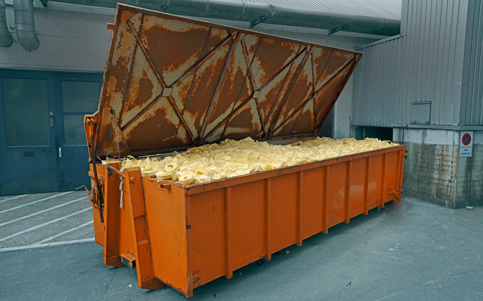 Een grote, oranje container met een inhoud van gele afvalstoffen staat tegen een muur, met een open deksel.